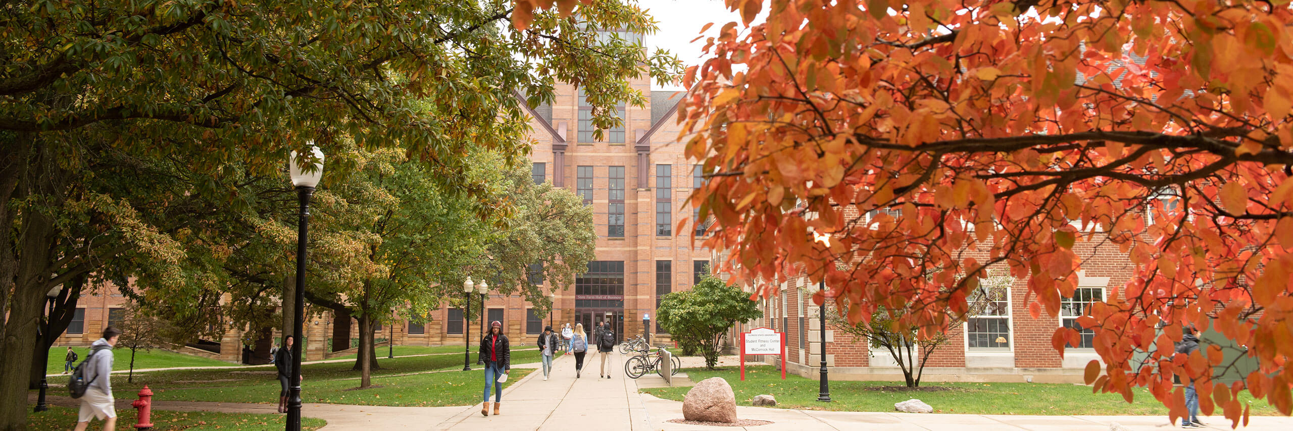ISU campus walkway with students, framed by green and orange autumn leaves and the State Farm Hall of Business building in the background.