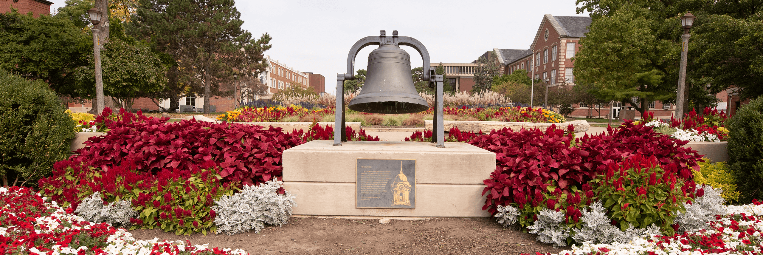 A large bell on a concrete pedestal surrounded by colorful flower beds and trees on a college campus.