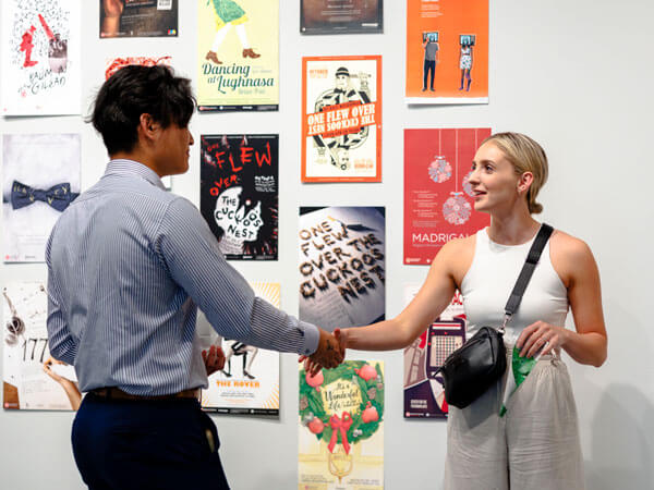 Two people shake hands in a gallery displaying graphic design pieces.